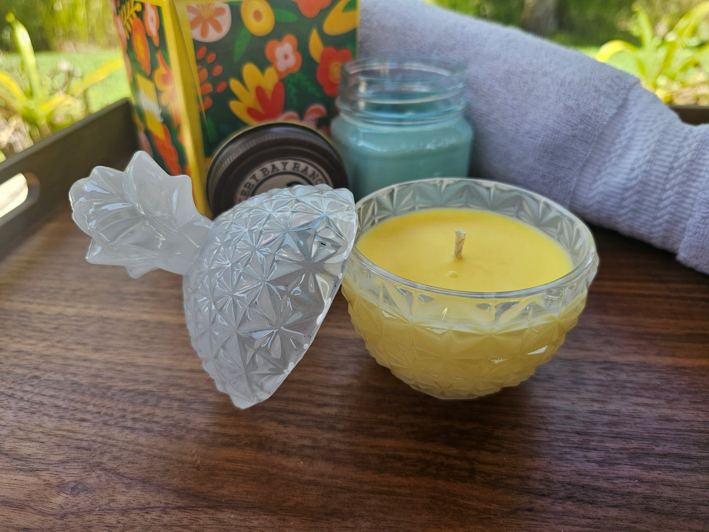 Yellow candle in a decorative pineapple glass holder on a wooden surface with a floral-patterned bag in the background.