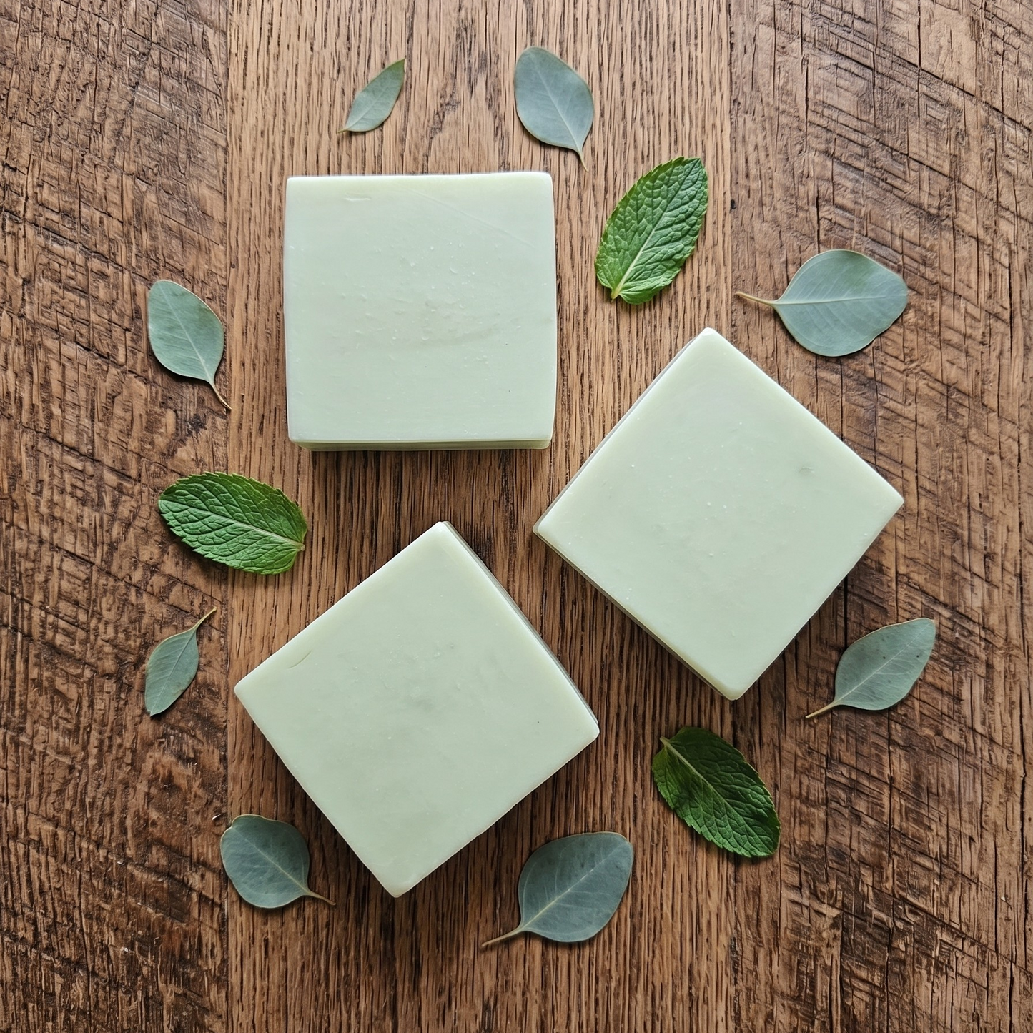 Three square bars of soap on a wooden surface with green leaves.